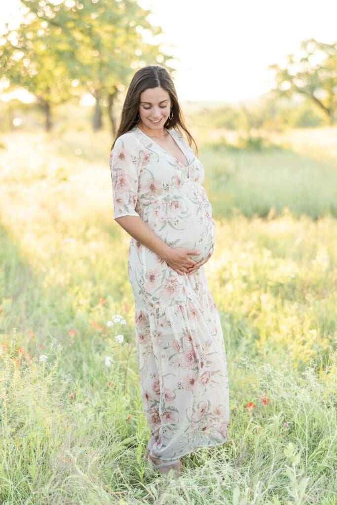 First time mom standing in a spring time open field holding her baby bump while looking down at it.