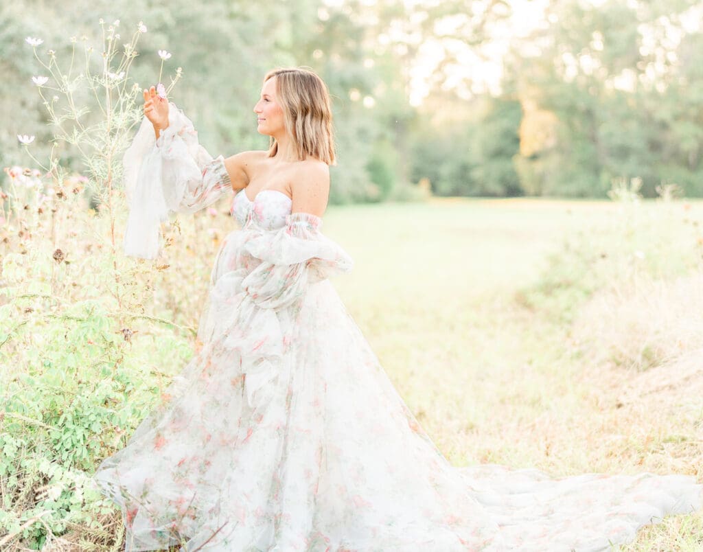 a first time mom wearing a white maternity gown with overlays of soft pastel florals featuring long puff sleeves. Designed by KH Fashions.