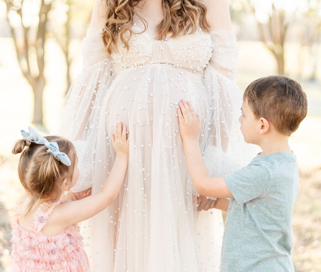 two siblings putting their hands on their mothers belly while holding their mothers hand.