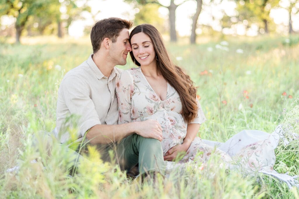 a maternity couple sitting in a field while husband nuzzles his face onto his wife. 